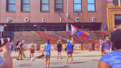 A time-lapse view of people with Pride flags throwing colorful streamers over a barricade outside a police station