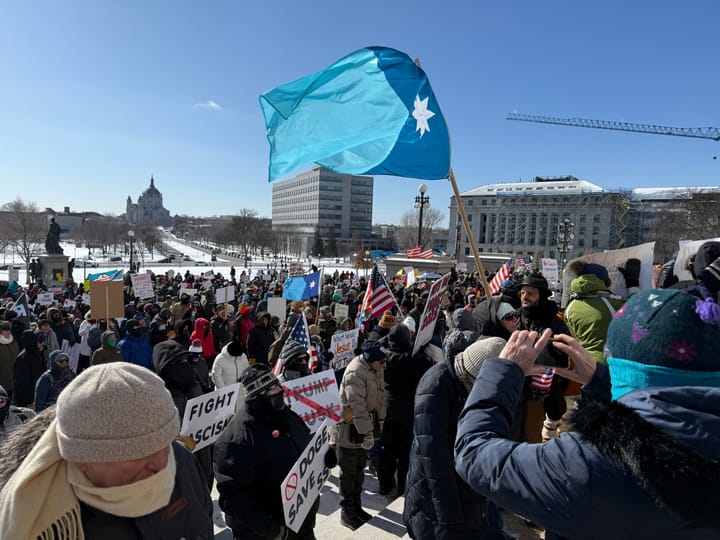 One of a crowd of demonstrators holds up the blue Minnesota flag, which snaps in the wind against the blue sky.