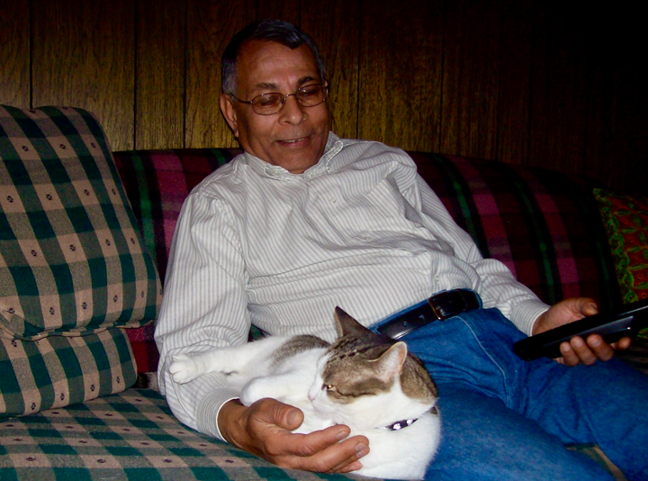 An Indian man in his late sixties sits on a sofa in jeans and a dress shirt, cuddled up next to a white-and-gray cat.